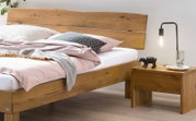Massives Holzbett mit breitem Kopfteil in natürlicher Eichenoptik, mit Bettwäsche dekoriert und Nachttisch mit Lampe daneben, fotografiert aus seitlicher Frontperspektive.