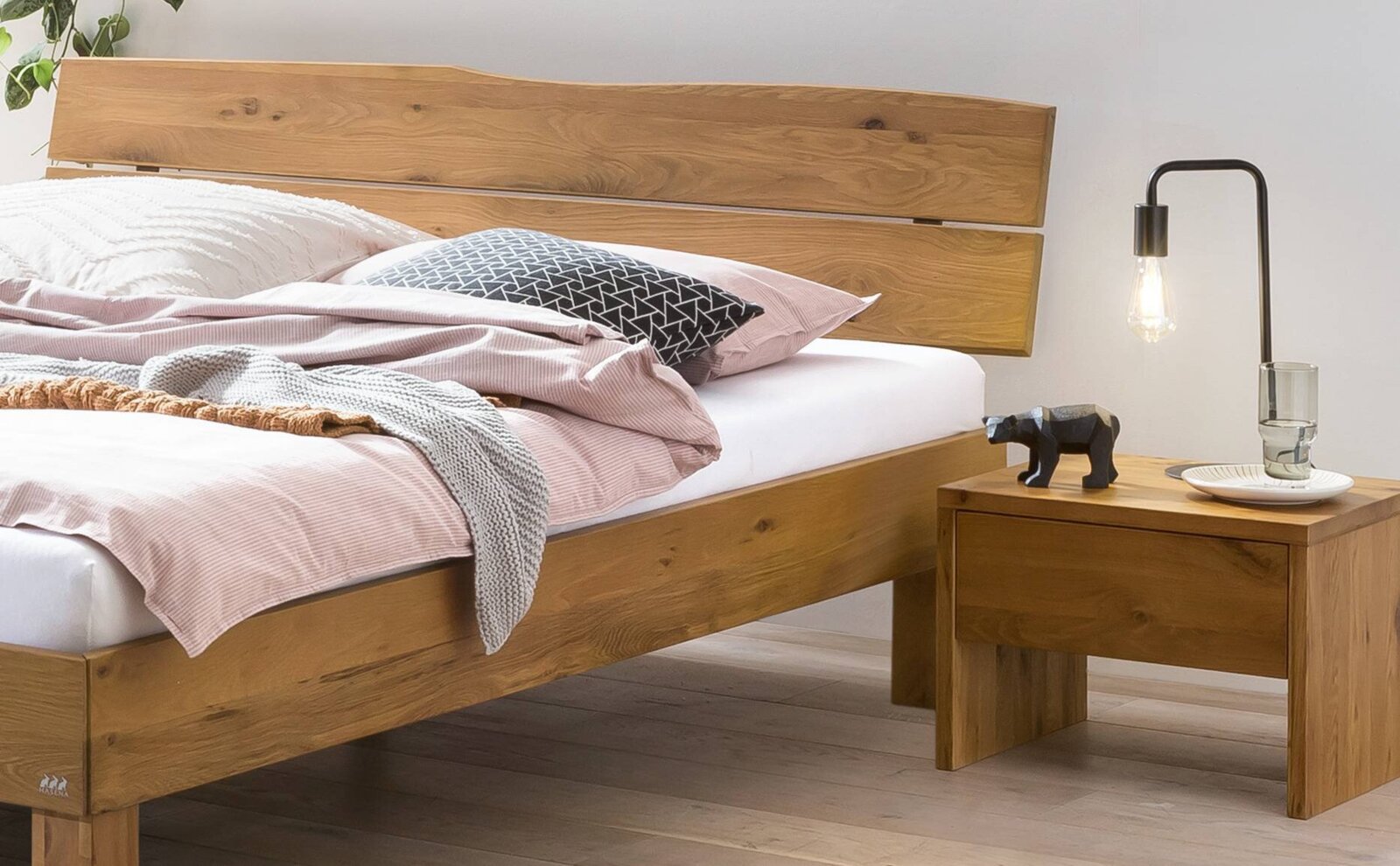 Massives Holzbett mit breitem Kopfteil in natürlicher Eichenoptik, mit Bettwäsche dekoriert und Nachttisch mit Lampe daneben, fotografiert aus seitlicher Frontperspektive.
