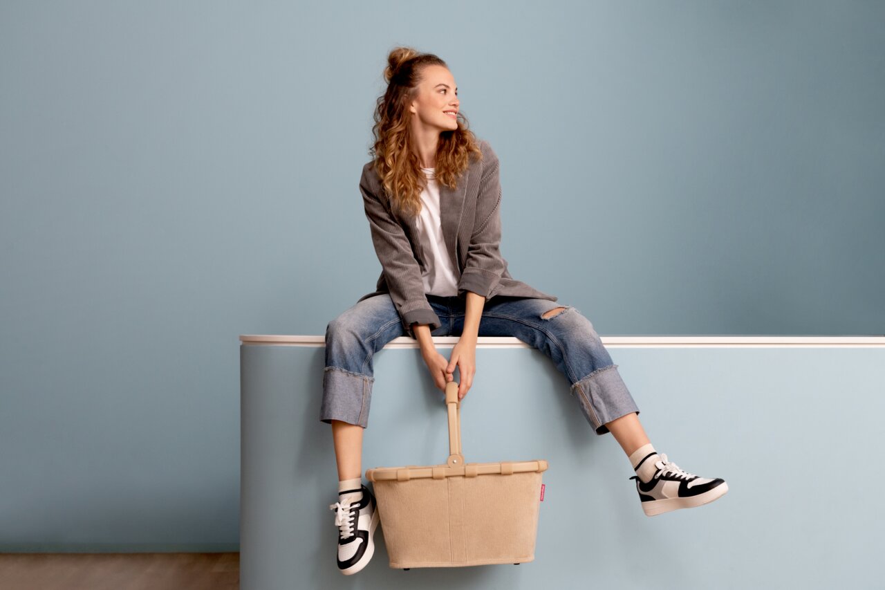 Frau sitzt lässig auf einer Kante und hält einen beigen Einkaufskorb mit Griff, seitliche Perspektive.