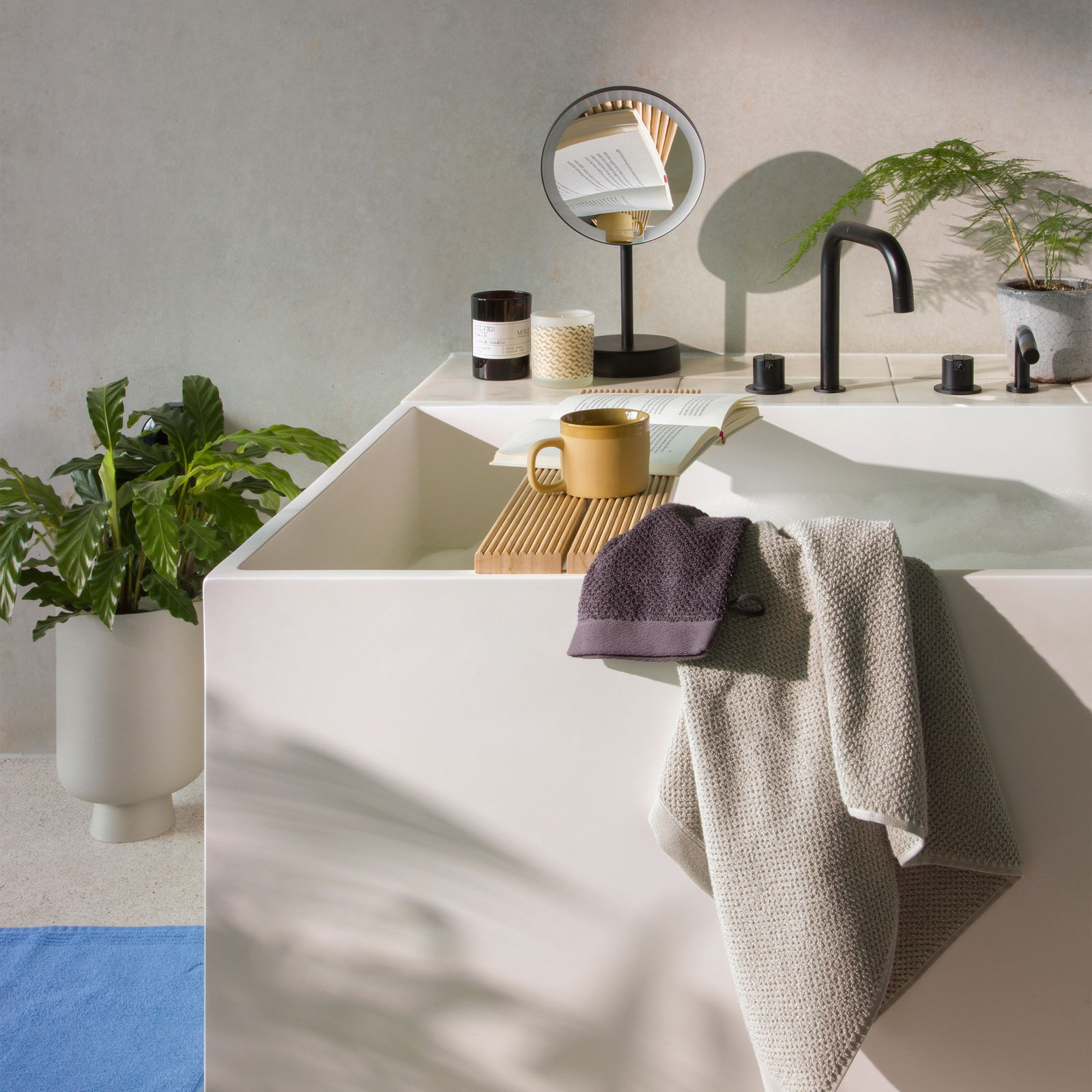 Modernes Badezimmer mit Handtuchset, bestehend aus einem grauen und einem dunklen Handtuch, drapiert über dem Rand einer weißen Badewanne. Im Hintergrund sind ein Spiegel, eine Tasse, ein Buch und Pflanzen zu sehen. Perspektive von der Seite der Badewanne