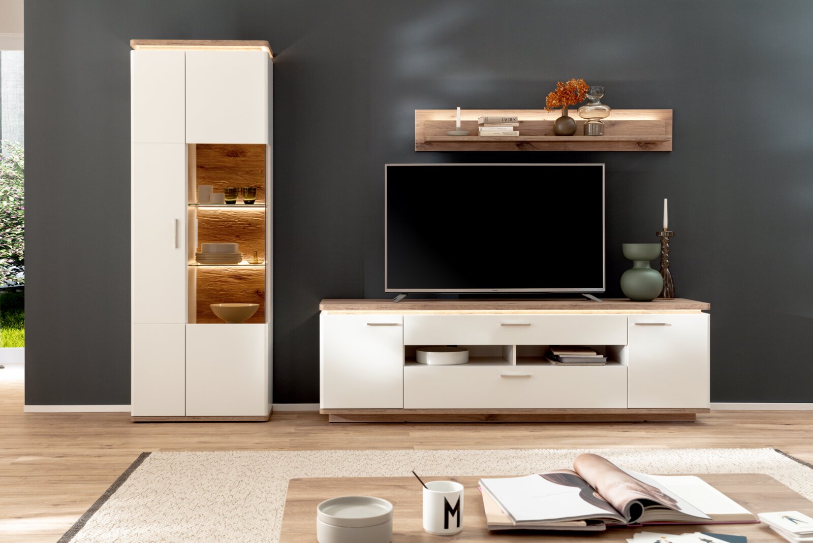 Moderne Wohnwand im Wohnzimmer: weiße TV-Kommode mit Holzplatte und Hängeregal sowie hohe Vitrine mit Holzrückwand und LED-Beleuchtung, frontale Ansicht auf dunkler Wand