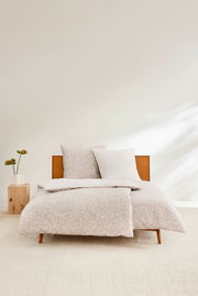 Satinbettwäsche-Set in Beige mit dezentem Muster auf einem Holzbett, frontal fotografiert, mit einem Nachttisch und einer Pflanze daneben.