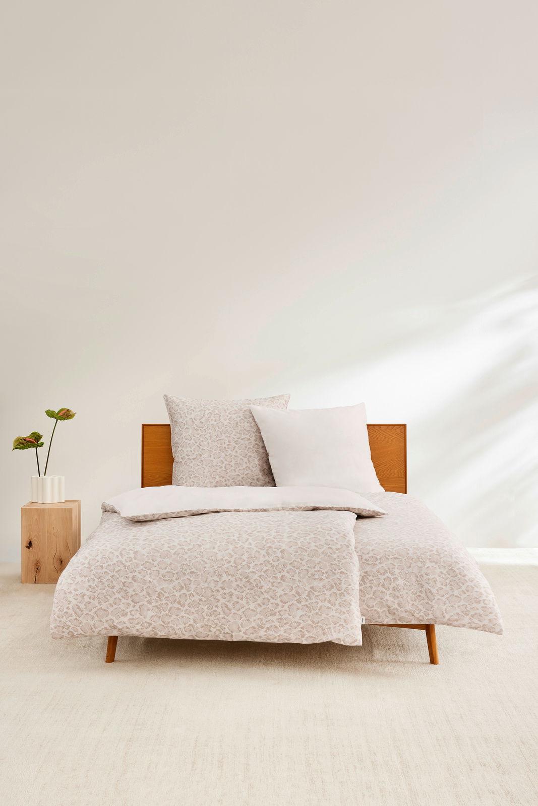Satinbettwäsche-Set in Beige mit dezentem Muster auf einem Holzbett, frontal fotografiert, mit einem Nachttisch und einer Pflanze daneben.