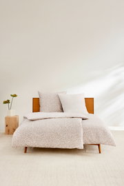 Satinbettwäsche-Set in Beige mit dezentem Muster auf einem Holzbett, frontal fotografiert, mit einem Nachttisch und einer Pflanze daneben.