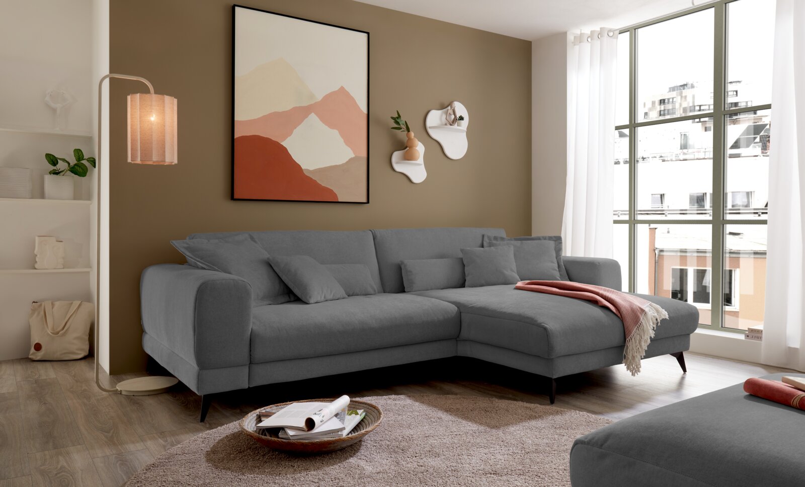 Set one by Musterring Ecksofa SET ONE MIDLAND Modernes graues L-förmiges Ecksofa mit Chaiselongue und Zierkissen im hellen Wohnzimmer, perspektivisch in schräger Vorderansicht von links aufgenommen.