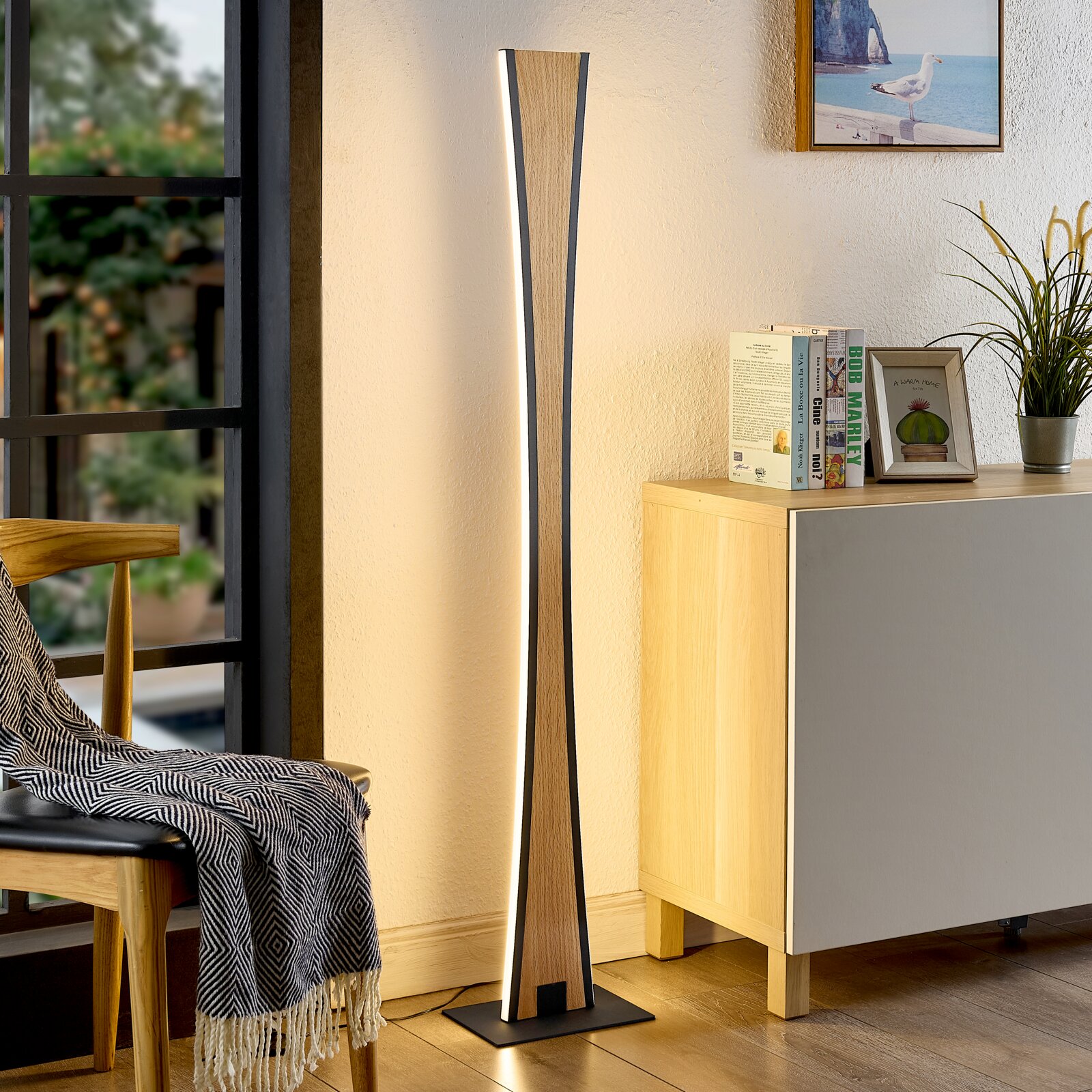 Schlanke LED-Stehlampe in Holzoptik mit schwarzem Rahmen und warmweißem indirektem Licht, freistehend auf quadratischem Sockel neben Sideboard im Wohnzimmer, frontale Perspektive.
