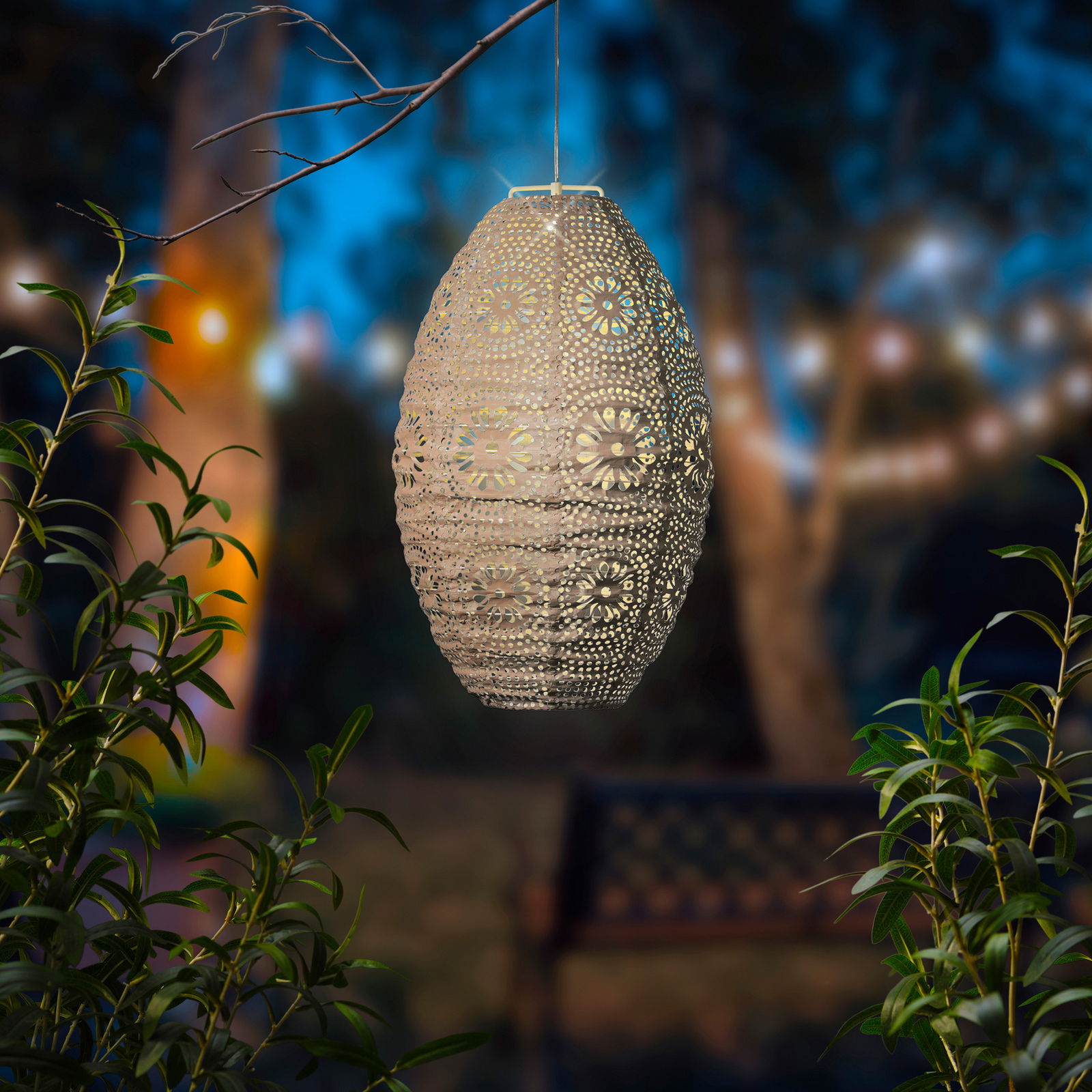 Hängende Solar Laterne in einem Garten bei Dämmerung, umgeben von Pflanzen, mit einem unscharfen Hintergrund aus Lichtern und Bäumen.