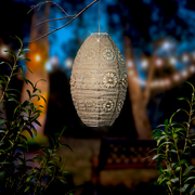 Hängende Solar Laterne in einem Garten bei Dämmerung, umgeben von Pflanzen, mit einem unscharfen Hintergrund aus Lichtern und Bäumen.