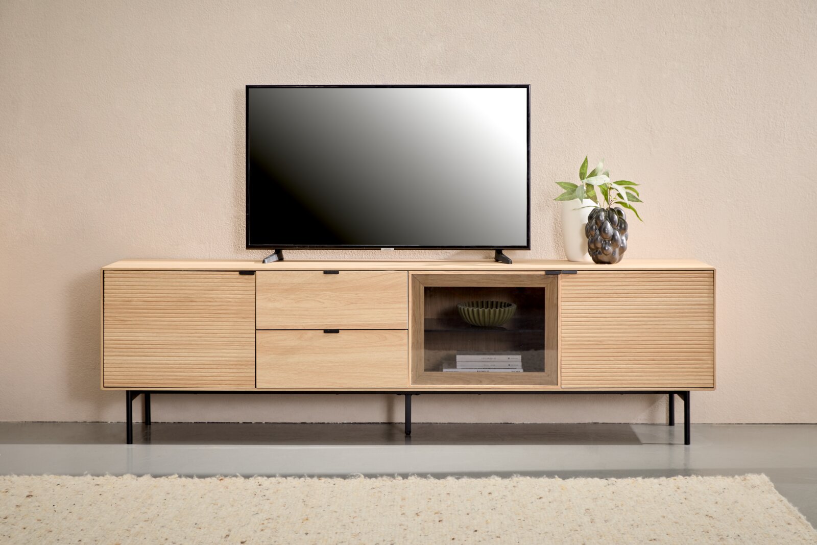 Modernes TV-Lowboard aus hellem Holz mit geriffelter Front, zwei Schubladen und Glasfach auf schwarzen Metallfüßen, frontal auf Augenhöhe fotografiert; darauf ein Fernseher und eine Vase mit Pflanze.