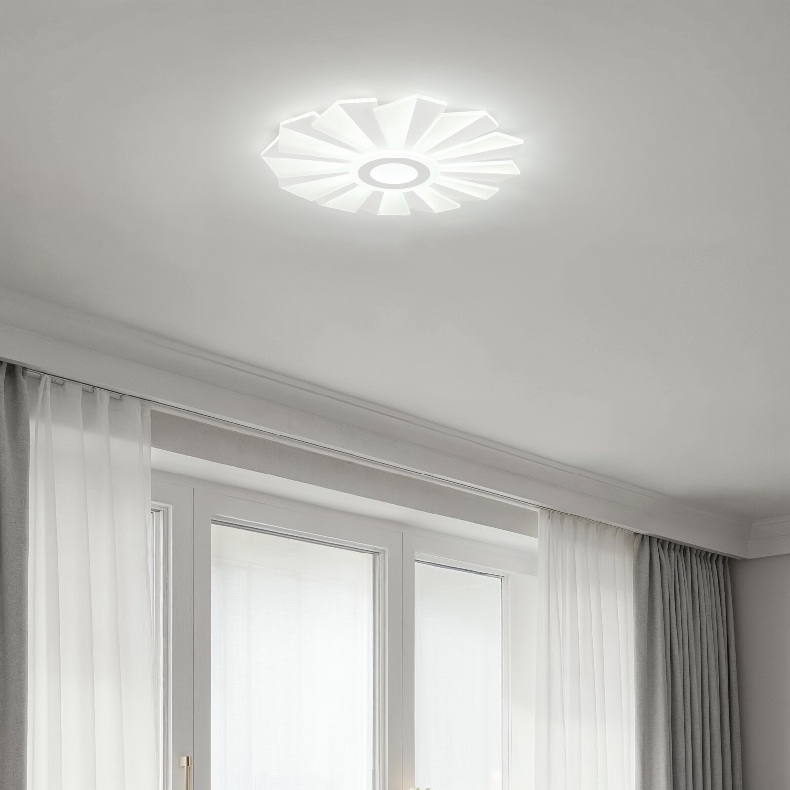 Moderne weiße LED-Deckenleuchte in Blumenform mit radialen Lamellen, eingeschaltet an der Zimmerdecke über Fenstern und Vorhängen, Aufnahme aus Untersicht nach oben