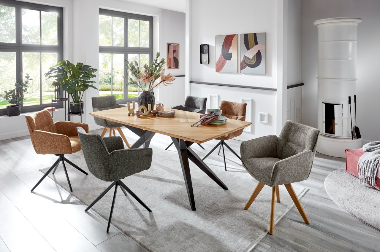 Moderner Esstisch mit Holzplatte und schwarzen Metallbeinen, umgeben von verschiedenen gepolsterten Stühlen in einem hellen Raum mit großen Fenstern. Perspektive von der Seite.