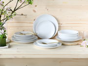 Ein 18-teiliges Tellerset aus weißem Porzellan, präsentiert auf einem Holztisch mit Blumen im Hintergrund. Die Perspektive zeigt das Set von vorne.