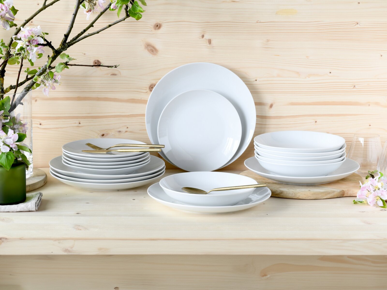 Ein 18-teiliges Tellerset aus weißem Porzellan, präsentiert auf einem Holztisch mit Blumen im Hintergrund. Die Perspektive zeigt das Set von vorne.