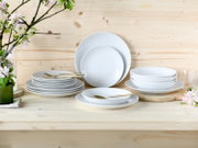 Ein 18-teiliges Tellerset aus weißem Porzellan, präsentiert auf einem Holztisch mit Blumen im Hintergrund. Die Perspektive zeigt das Set von vorne.