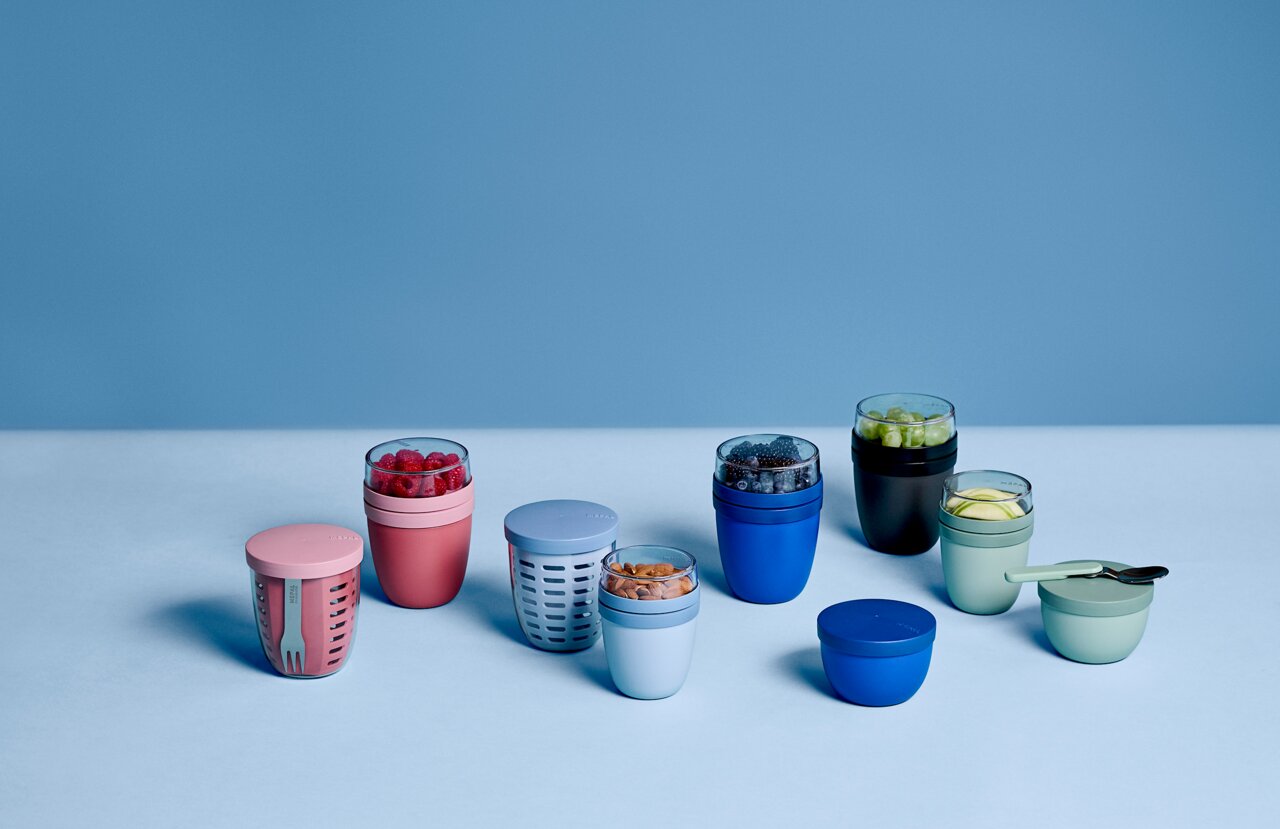 Verschiedene bunte Fruchtbehälter in elliptischer Form, gefüllt mit Obst und Nüssen, auf einer blauen Oberfläche aus der Vogelperspektive.