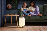 Akku-Leuchte und Lautsprecher in einem modernen Wohnzimmer, auf dem Boden neben einem Tisch stehend, mit zwei Kindern auf einem Sofa im Hintergrund. Perspektive von vorne.