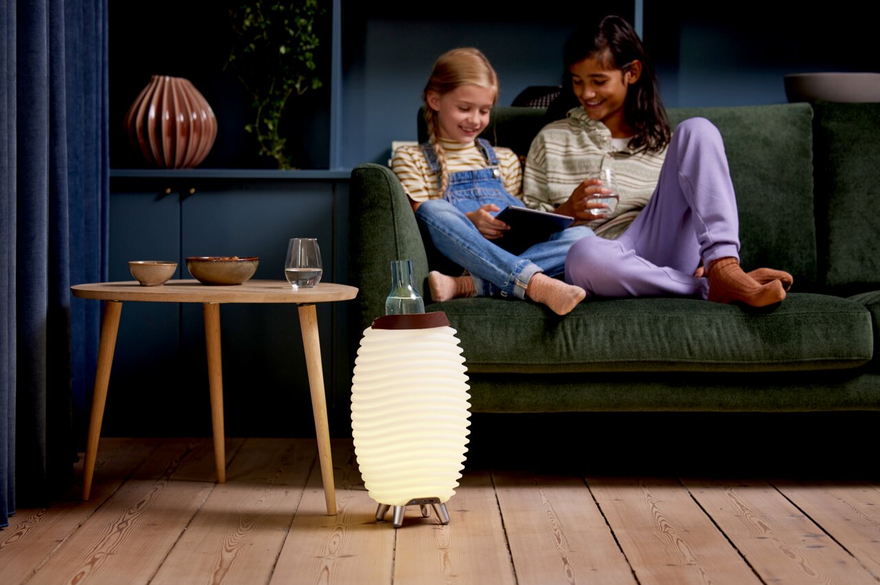 Een acculamp en luidspreker in een moderne woonkamer, op de grond naast een tafel, met twee kinderen op een bank op de achtergrond. Aanzicht van voren.