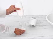 Nahaufnahme eines Seifenspenders auf einer Marmoroberfläche, während eine Person Seife in ein Glasgefäß füllt.