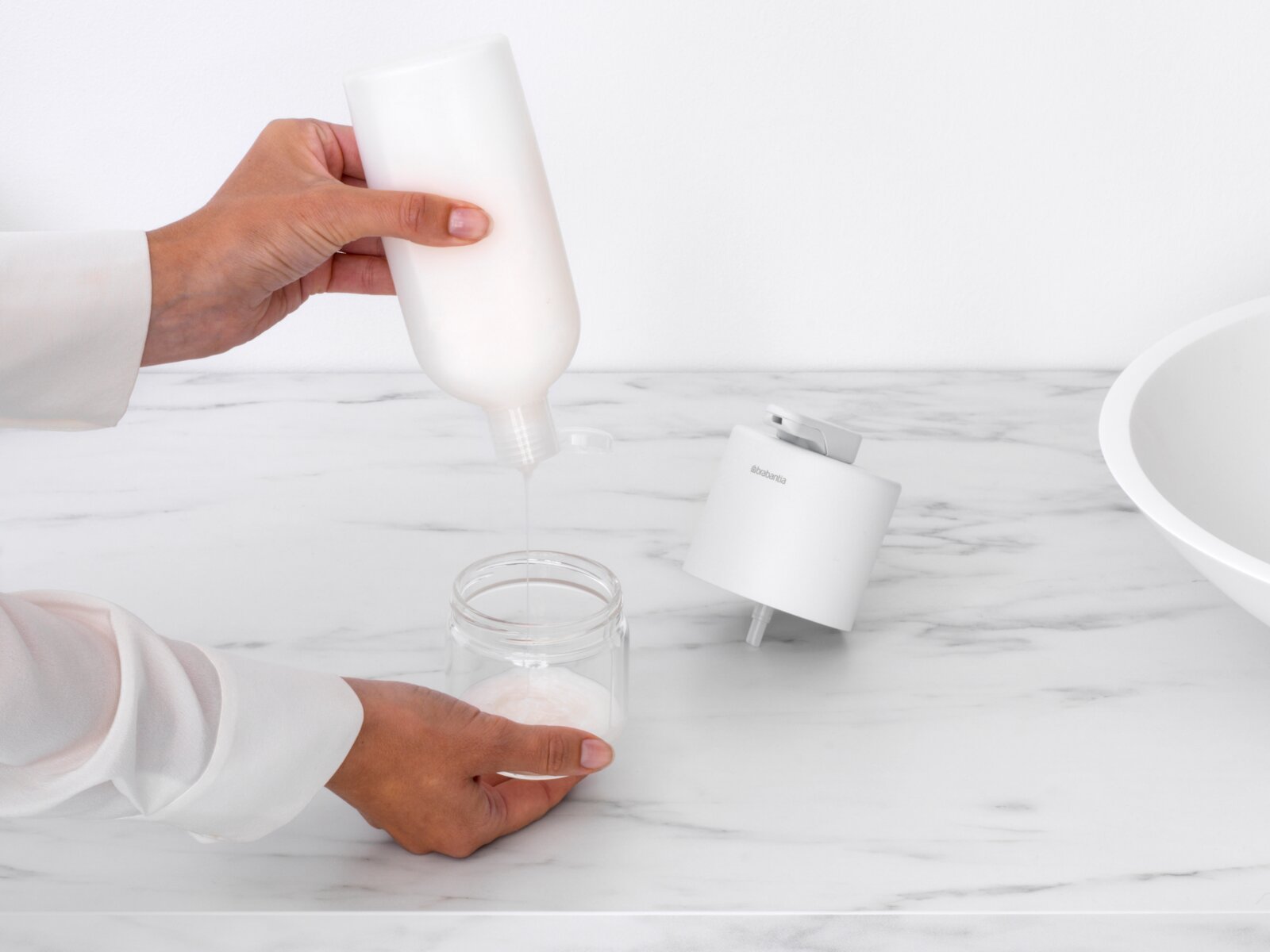 Close-up van een zeepdispenser op een marmeren oppervlak, terwijl iemand zeep in een glazen pot giet.
