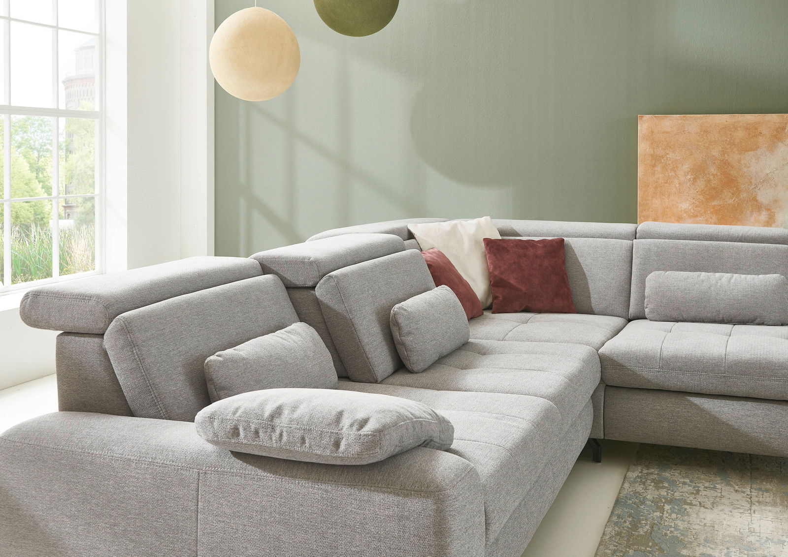 Graue Eckgarnitur mit verstellbaren Kopfstützen und mehreren Kissen in einem hellen Raum mit großen Fenstern, seitliche Perspektive.