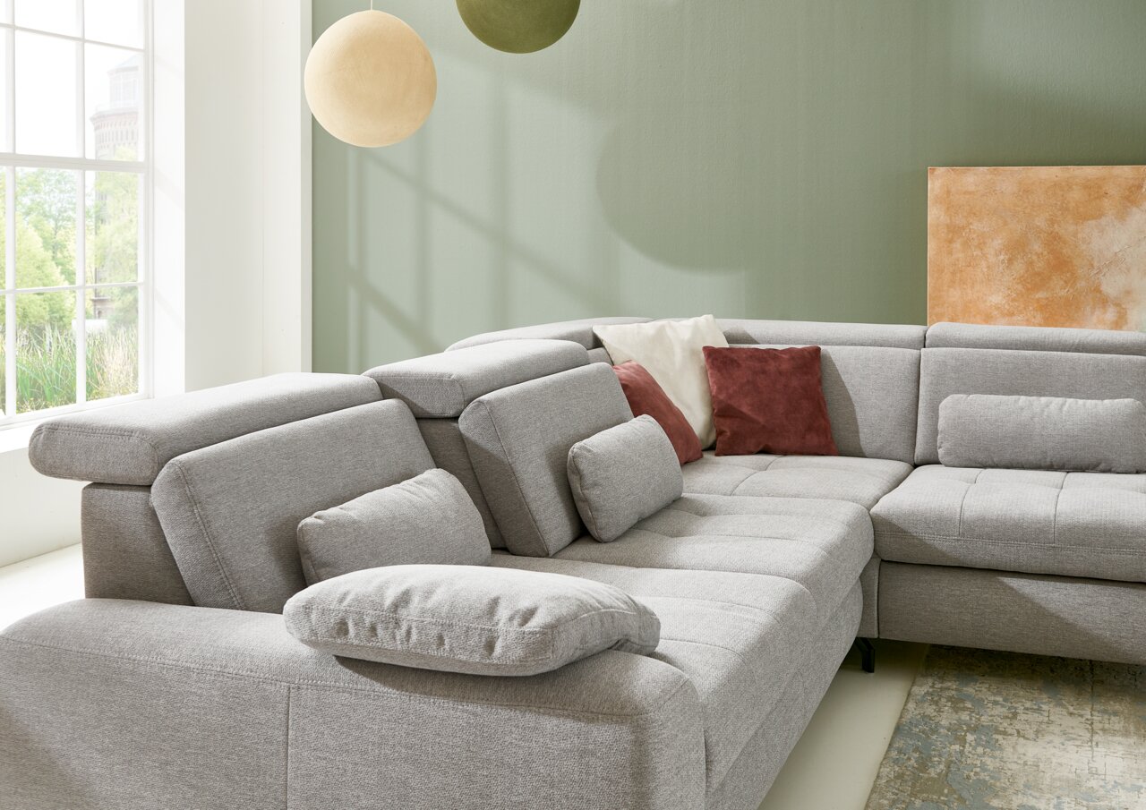 Graue Eckgarnitur mit verstellbaren Kopfstützen und mehreren Kissen in einem hellen Raum mit großen Fenstern, seitliche Perspektive.