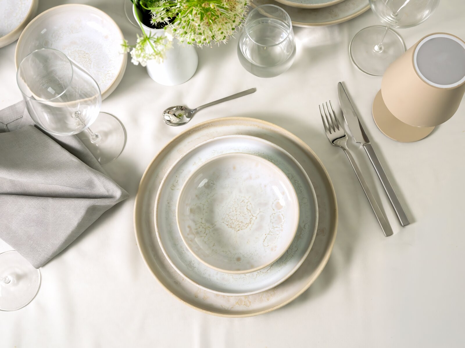 Draufsicht auf eine elegant gedeckte Festtafel mit cremefarbenem Geschirr in Craquelé-Optik, Besteck, Gläsern, grauer Stoffserviette und weißen Blüten
