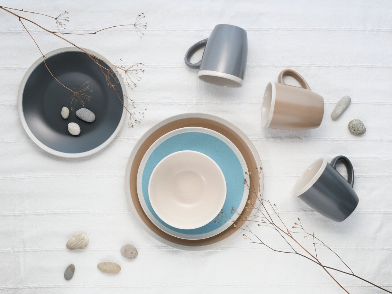 Vogelperspektive auf ein stilvolles Kombiservice-Set 'Nordic Living' mit Tellern und Tassen in gedeckten Farben auf einem weißen Tuch, dekoriert mit Steinen und Zweigen.