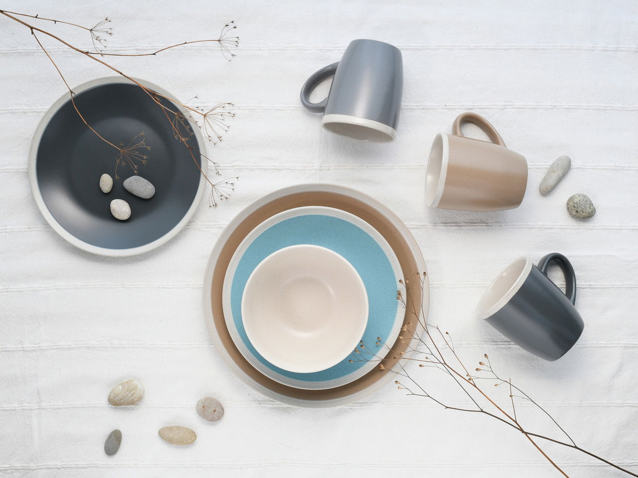 Vogelperspektive auf ein stilvolles Kombiservice-Set 'Nordic Living' mit Tellern und Tassen in gedeckten Farben auf einem weißen Tuch, dekoriert mit Steinen und Zweigen.