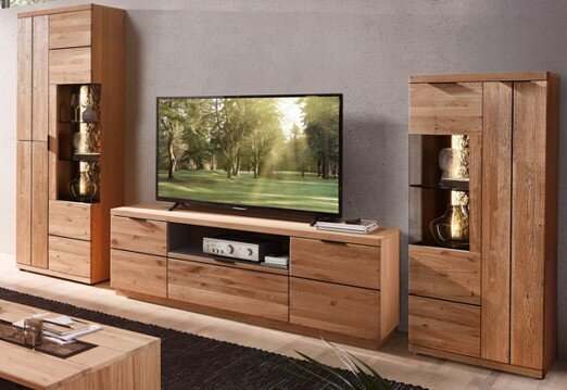 Wohnwand aus massivem Holz im Wohnzimmer: TV-Lowboard mit Schubladen und offenem Fach, flankiert von zwei hohen Vitrinenschränken mit Glasböden und Beleuchtung, frontale Perspektive