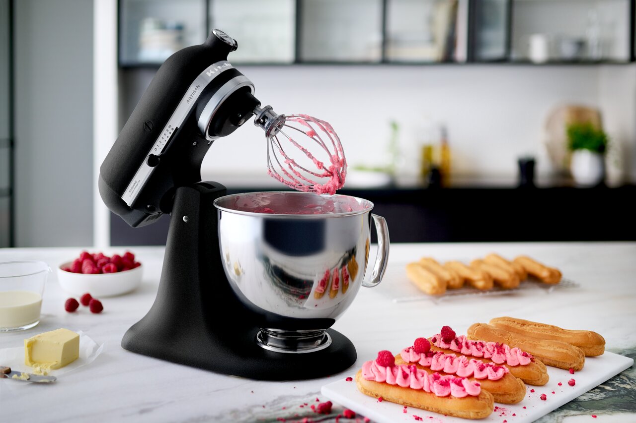 Schwarze Küchenmaschine mit Rührbesen und Edelstahlschüssel, gefüllt mit rosa Teig, auf einer Küchenarbeitsplatte. Im Hintergrund sind Himbeeren, Sahne und Gebäck zu sehen. Perspektive von vorne links.