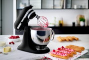 Schwarze Küchenmaschine mit Rührbesen und Edelstahlschüssel, gefüllt mit rosa Teig, auf einer Küchenarbeitsplatte. Im Hintergrund sind Himbeeren, Sahne und Gebäck zu sehen. Perspektive von vorne links.
