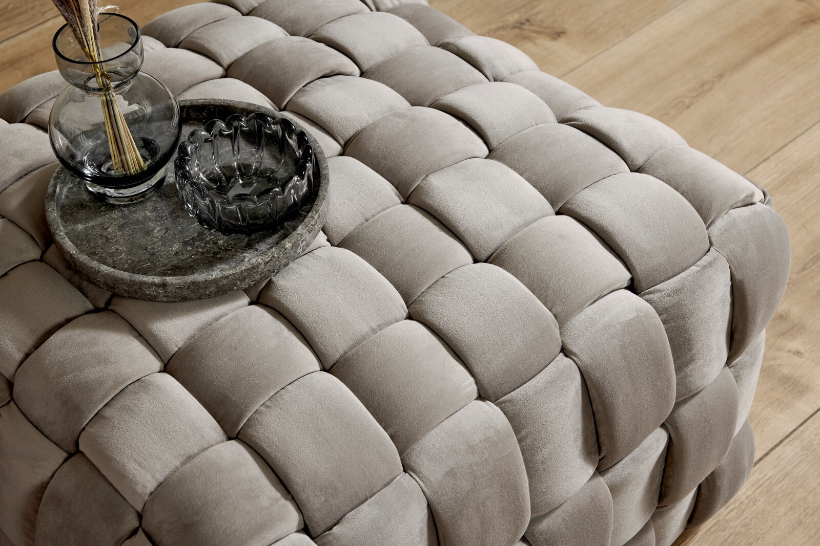 Nahaufnahme eines gepolsterten Hockers mit geflochtenem Design in Beige, darauf ein runder Steintablett mit Glasvasen, aus der Vogelperspektive.