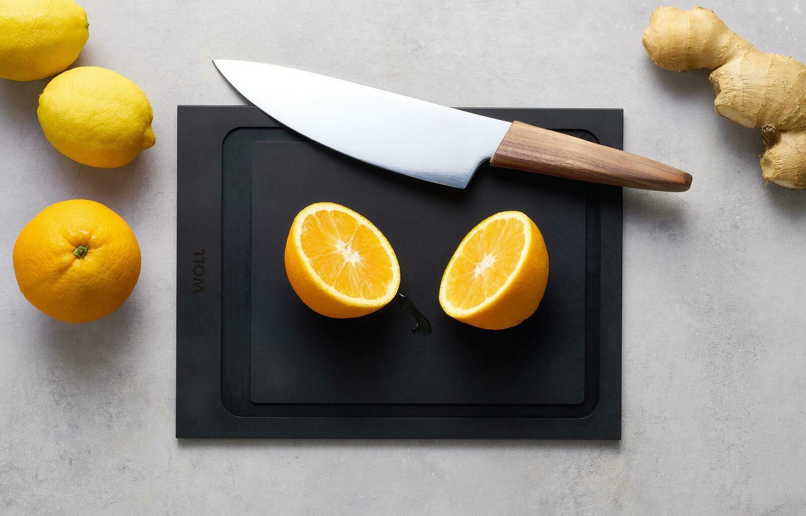 Woll Schneidebrett CULT & SERVE Schneidebrett M 30x22cm mit einem Messer und einer halbierten Orange, umgeben von Zitronen und Ingwer, aus der Vogelperspektive.