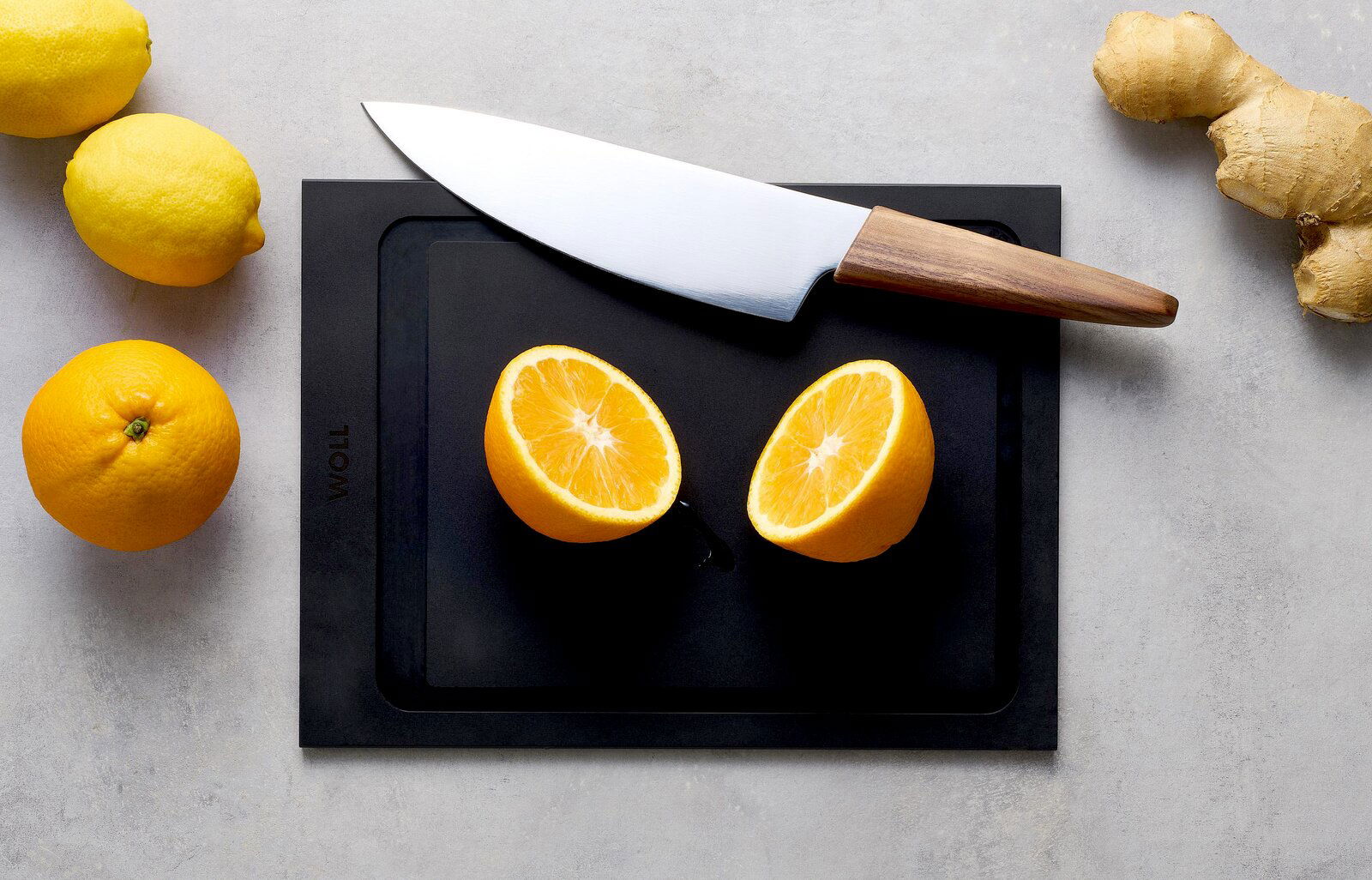 Woll Schneidebrett CULT & SERVE Schneidebrett M 30x22cm mit einem Messer und einer halbierten Orange, umgeben von Zitronen und Ingwer, aus der Vogelperspektive.