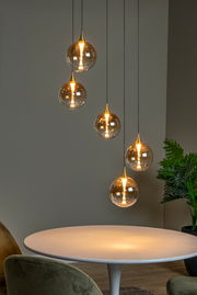 Moderne Hängeleuchte mit fünf kugelförmigen, goldenen Lampenschirmen, die in unterschiedlichen Höhen über einem runden Tisch hängen. Seitliche Perspektive.