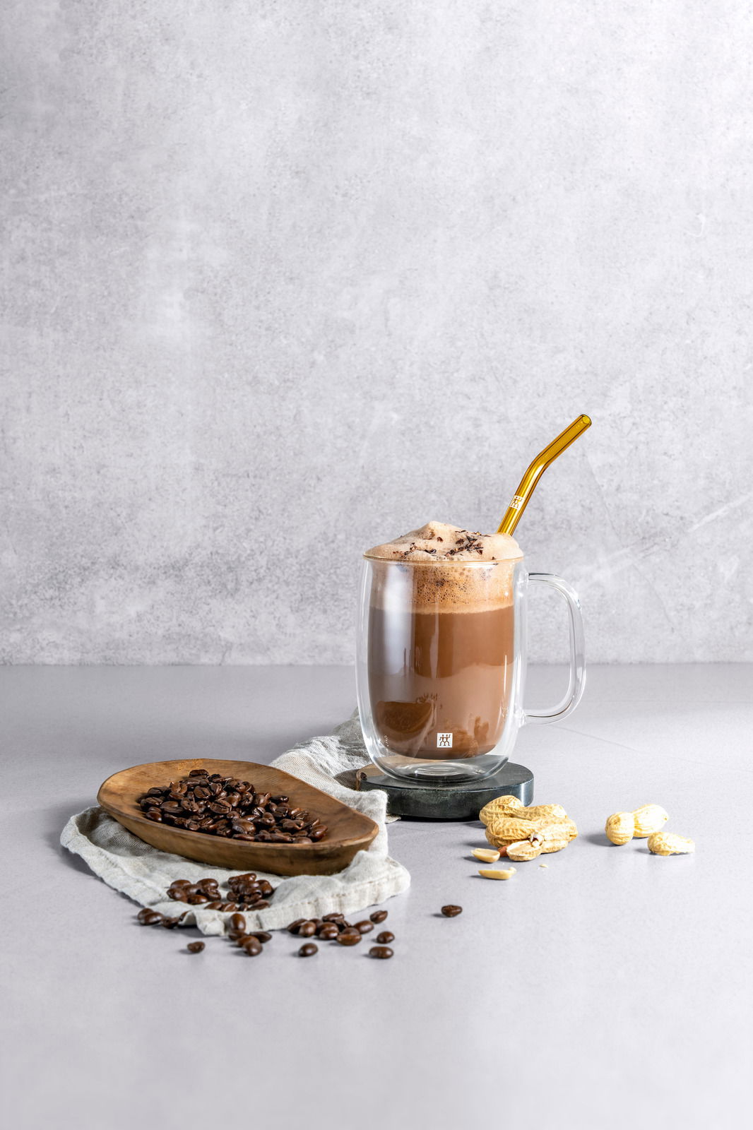 Latte Macchiato in einem doppelwandigen Glas mit goldenem Strohhalm, umgeben von Kaffeebohnen und Erdnüssen, seitliche Perspektive.