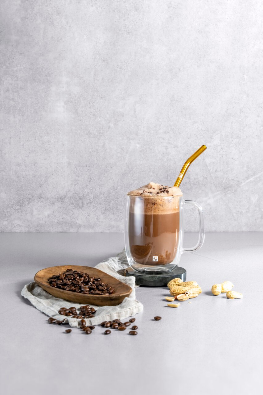 Latte macchiato in een dubbelwandig glas met een gouden rietje, omringd door koffiebonen en pinda’s, zijaanzicht.