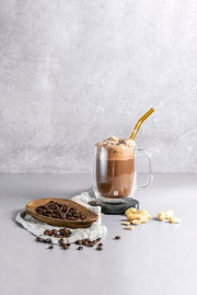Latte macchiato in een dubbelwandig glas met een gouden rietje, omringd door koffiebonen en pinda’s, zijaanzicht.