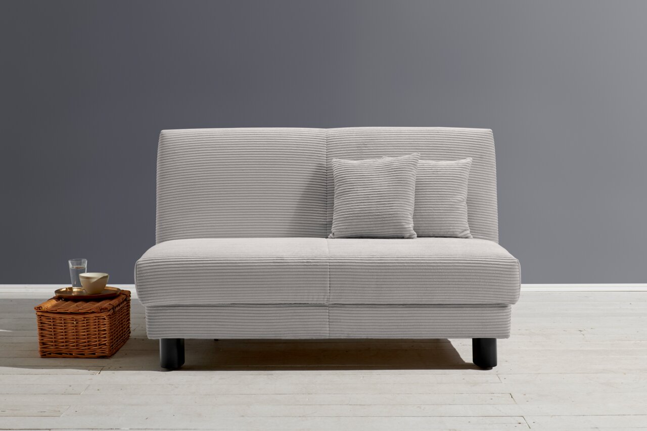 Z2 Schlafsofa ENNY Graues Funktionssofa mit zwei passenden Kissen, frontal fotografiert, auf hellem Holzboden vor grauer Wand, neben einem Korbtisch mit Glas und Schale.