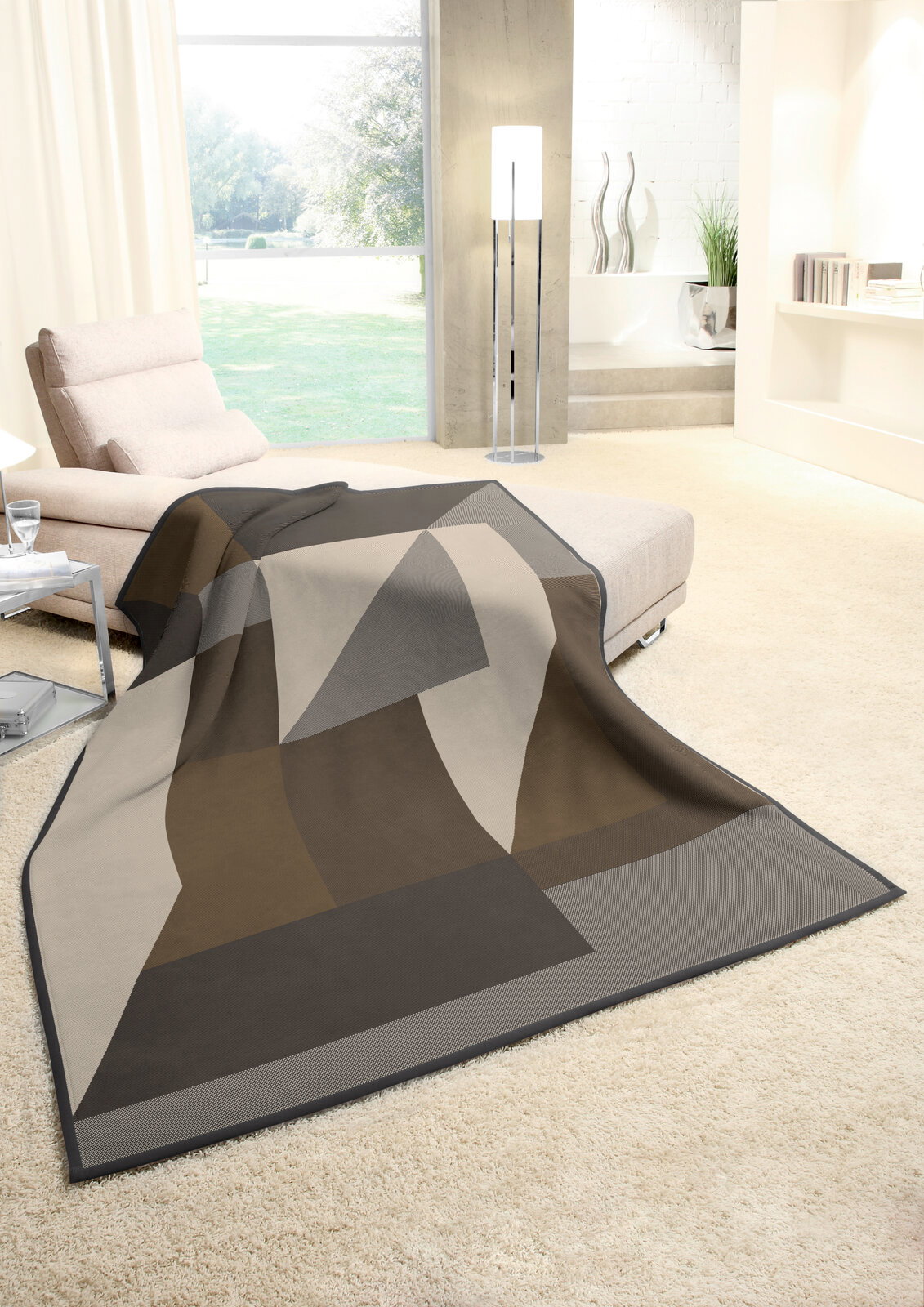 Moderne Wohndecke 150x200cm in geometrischem Design, drapiert auf einem Sofa in einem hellen Wohnzimmer mit Blick auf den Garten.