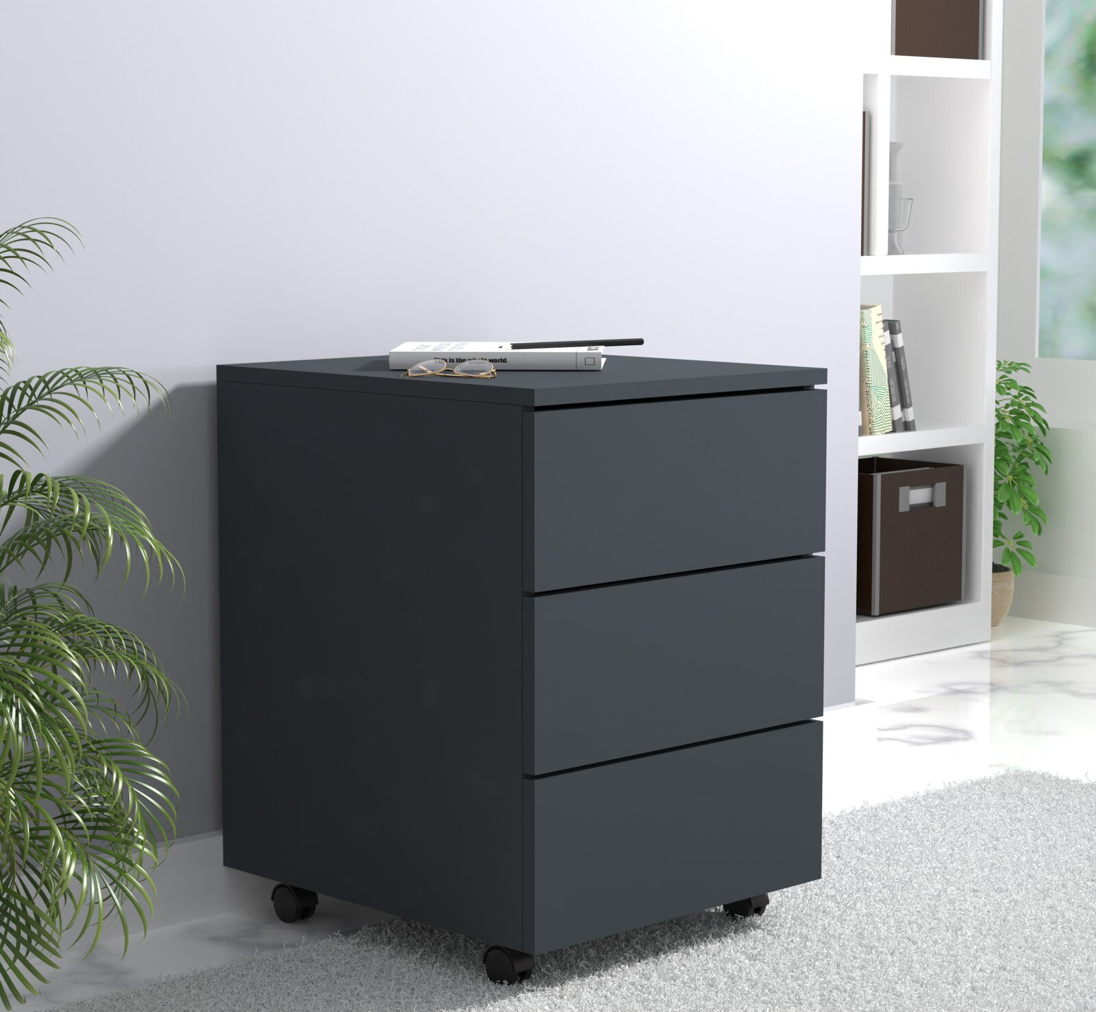 Dunkelgrauer, moderner Rollcontainer mit drei Schubladen auf Rollen, schräg von vorne links in einem hellen Büro/Wohnraum fotografiert auf der Ablage liegen Buch, Stift und Brille.