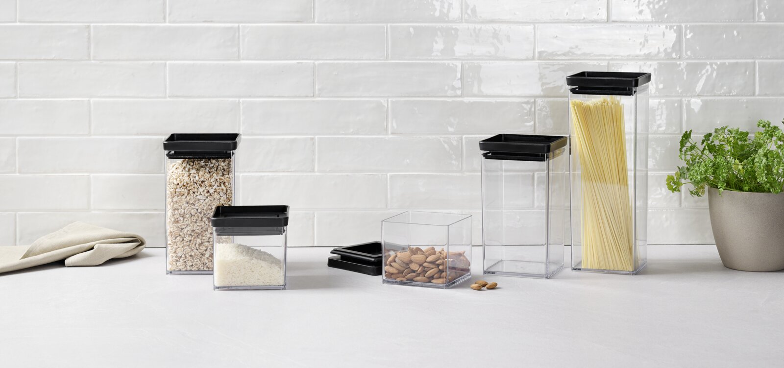 Homeware Set van 5 voorraadbussen Set van 5 voorraadbussen van doorzichtig kunststof met zwarte deksels, gevuld met diverse levensmiddelen, op een keukenblad voor een witte tegelwand, zijaanzicht.