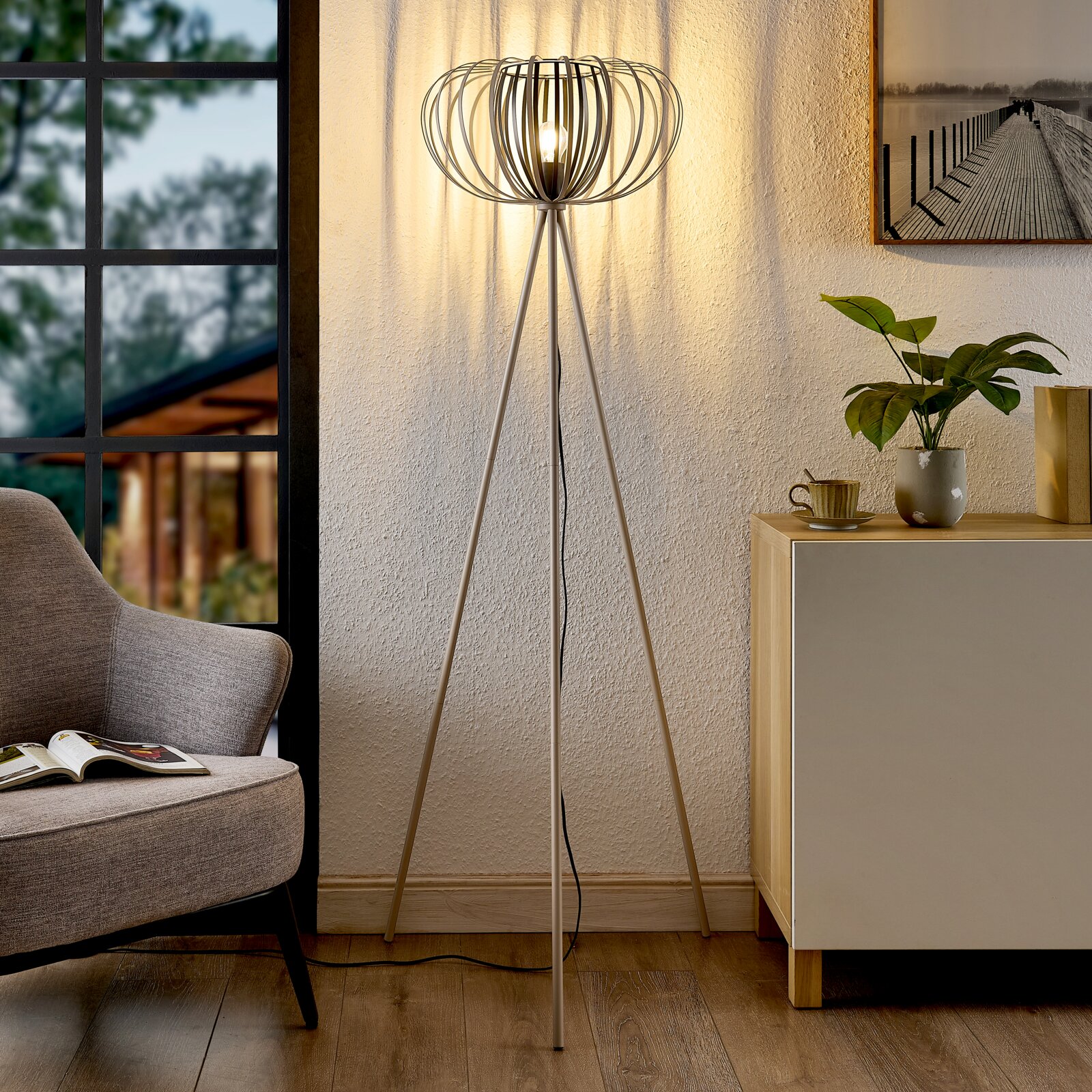Novel Staande lamp STEHLEUCHTE Moderne staande lamp met driepoot en decoratieve lampenkap in een gezellige woonkamer, gezien vanaf de zijkant.