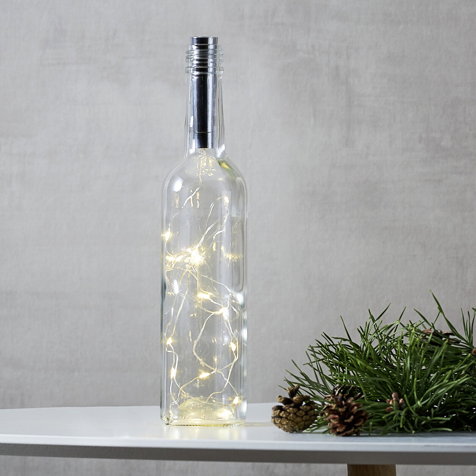 STAR TRADING LED-Lichterkette für Flaschen DEW DROP TUBE Transparente Glasflasche mit warmweißer LED-Lichterkette im Inneren auf weißem Tisch, rechts Tannenzweig mit Zapfen, frontal aus Augenhöhe fotografiert.