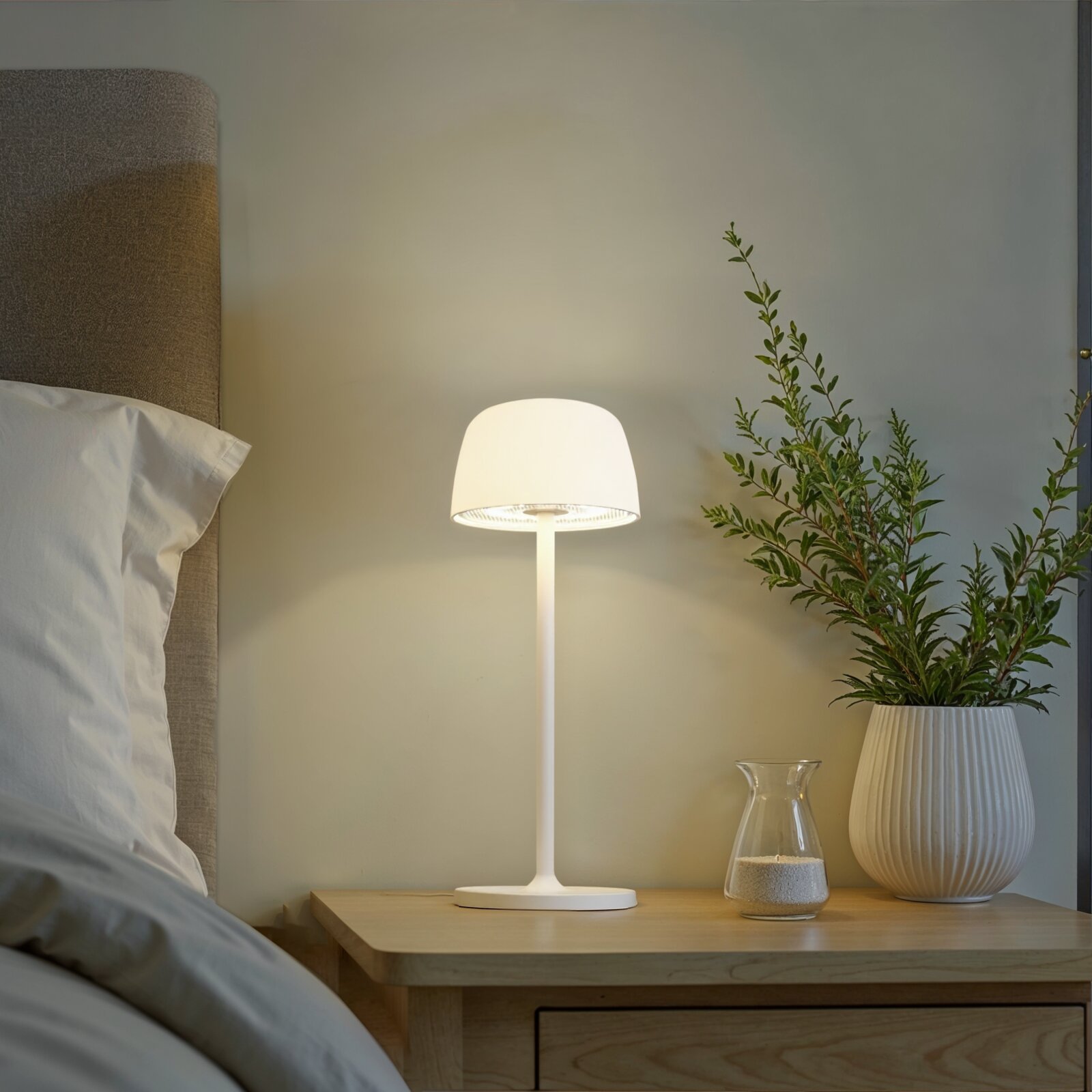Weiße moderne Tischlampe mit rundem Schirm auf einem Holz-Nachttisch neben Bett und Pflanze, Frontansicht auf Augenhöhe