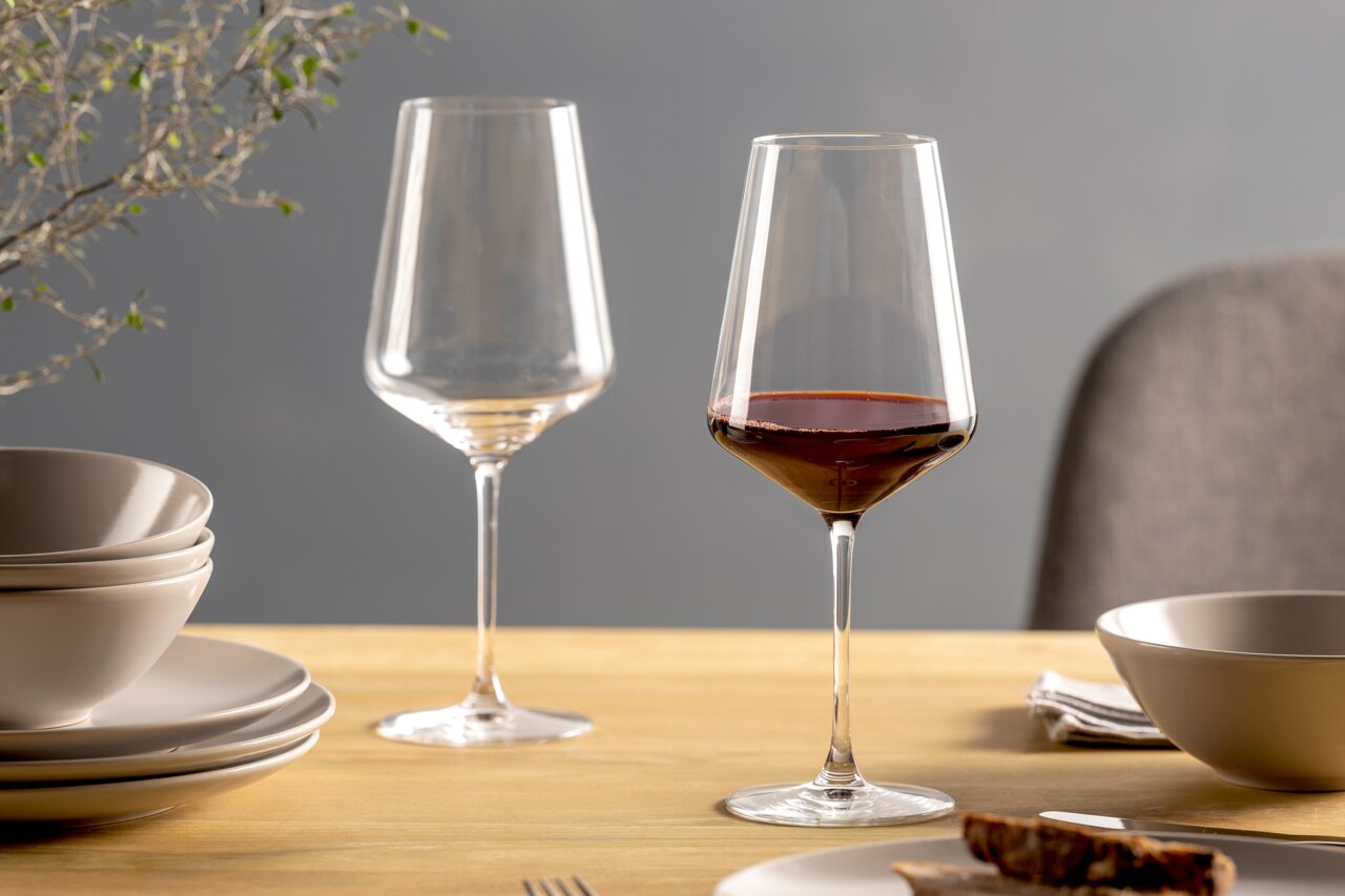 Zwei Rotweingläser auf einem gedeckten Tisch, eines gefüllt mit Rotwein, aus seitlicher Perspektive fotografiert.