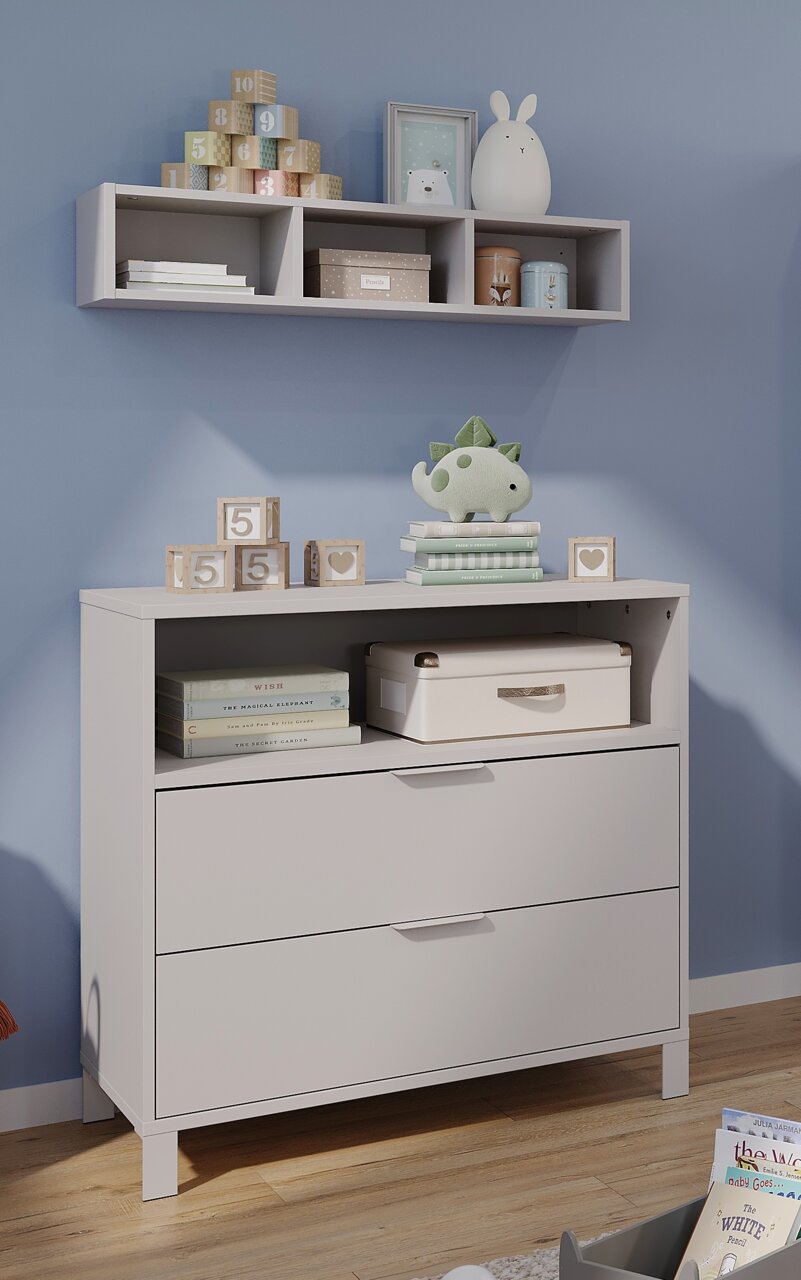 Kommode und Wandregal in einem Babyzimmer, frontal fotografiert. Auf der Kommode stehen Spielzeug und Bücher, während das Regal mit Dekorationen und Aufbewahrungsboxen gefüllt ist.