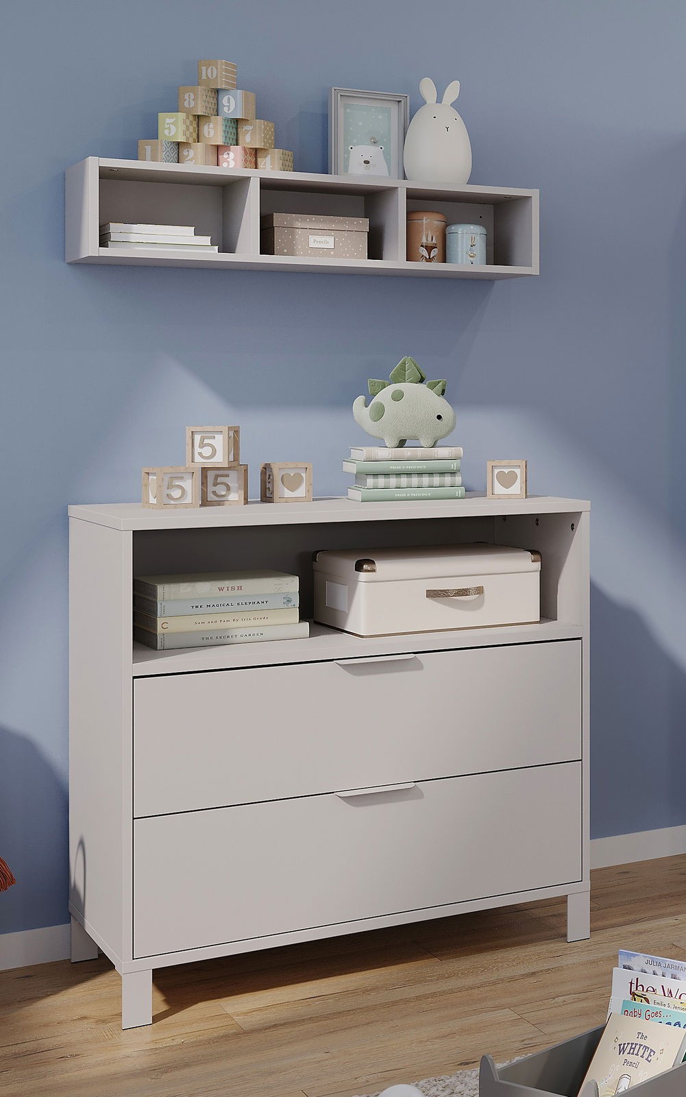 Kommode und Wandregal in einem Babyzimmer, frontal fotografiert. Auf der Kommode stehen Spielzeug und Bücher, während das Regal mit Dekorationen und Aufbewahrungsboxen gefüllt ist.