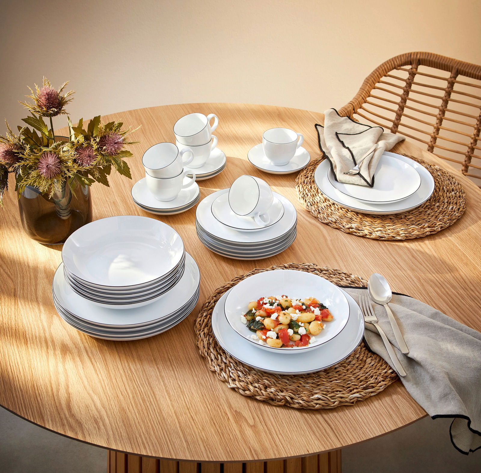 Elegantes Kombiservice Lido Black 30-teilig auf einem Holztisch, bestehend aus Tellern, Tassen und Schalen, mit einer modernen Tischdekoration aus Blumen und geflochtenen Platzsets, aus der Vogelperspektive fotografiert.