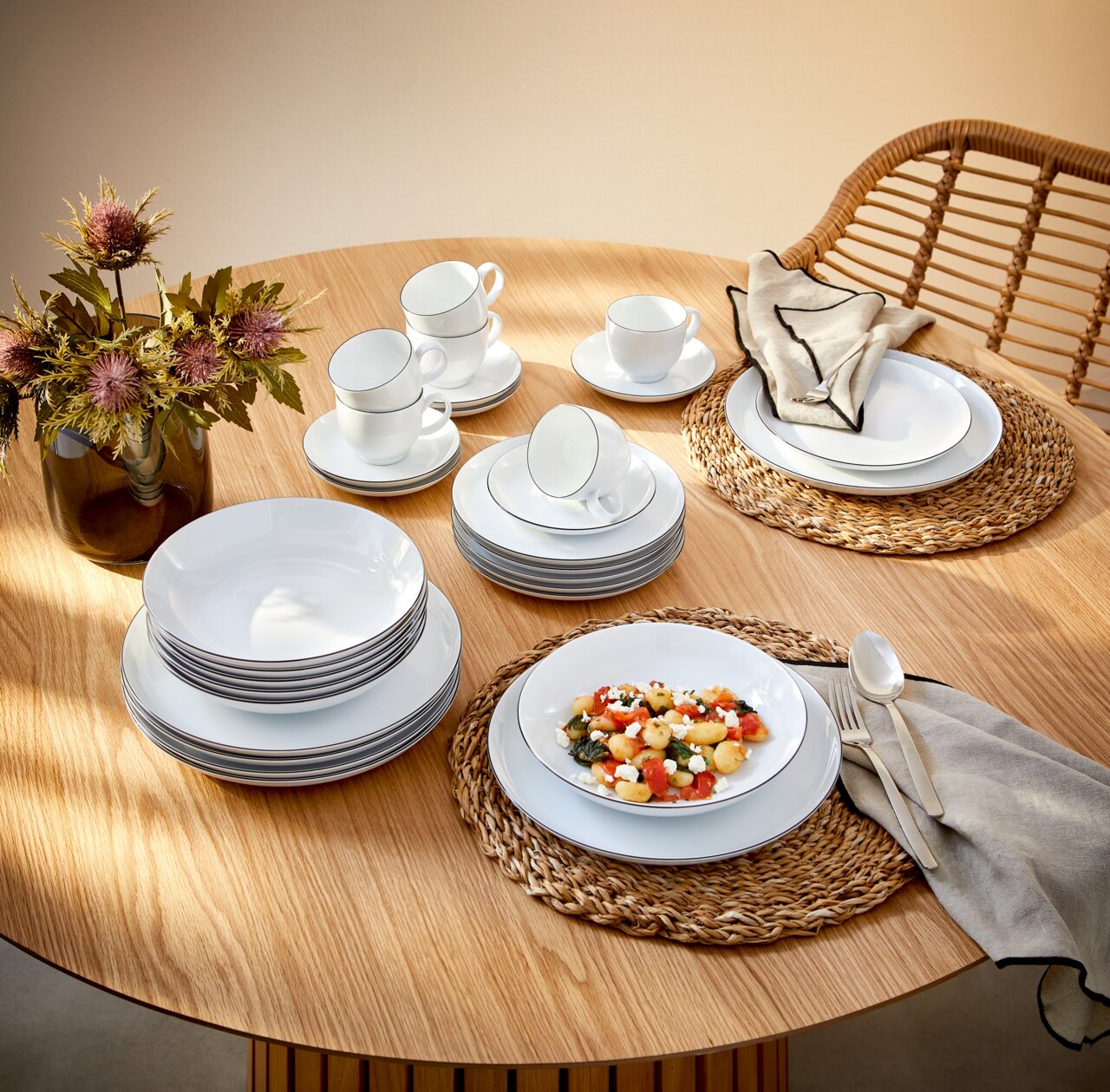 Elegantes Kombiservice Lido Black 30-teilig auf einem Holztisch, bestehend aus Tellern, Tassen und Schalen, mit einer modernen Tischdekoration aus Blumen und geflochtenen Platzsets, aus der Vogelperspektive fotografiert.